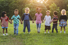 A diverse group of children holding hands and walking in the grass with sunshine behind them.