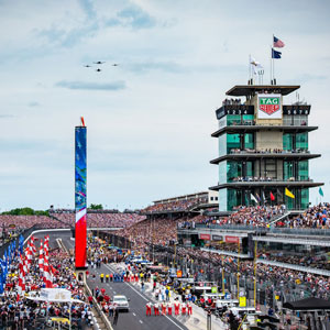 Indianapolis 500 race  showing the crowds in the stands and the Pagota with four airforce fighter jets in the sky flying above the motor speedway track.